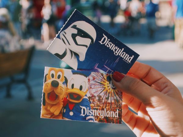 Two Disneyland tickets are being held up, one featuring a stormtrooper and the other showcasing Goofy, Donald Duck, and a Ferris wheel.