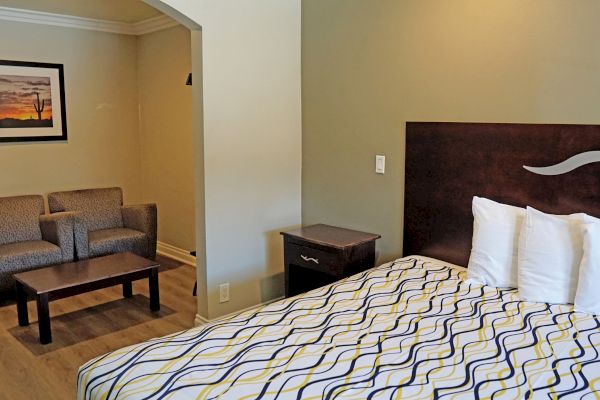 The image shows a hotel room with a bed, two chairs, a small table, and a framed picture on the wall. The decor is simple and modern.