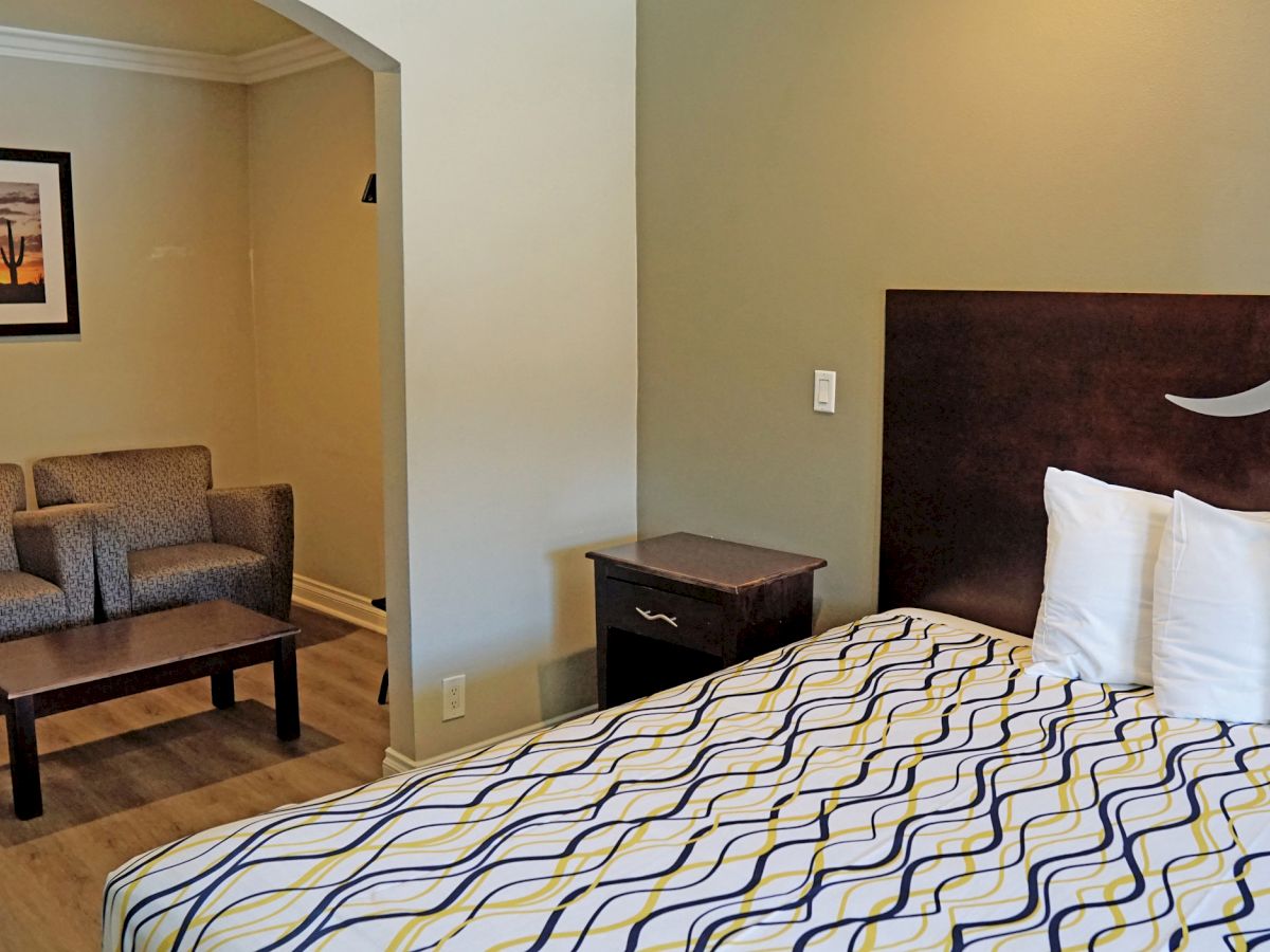 The image shows a hotel room with a bed, bedside table, a seating area with two chairs and a coffee table, and a framed picture on the wall.