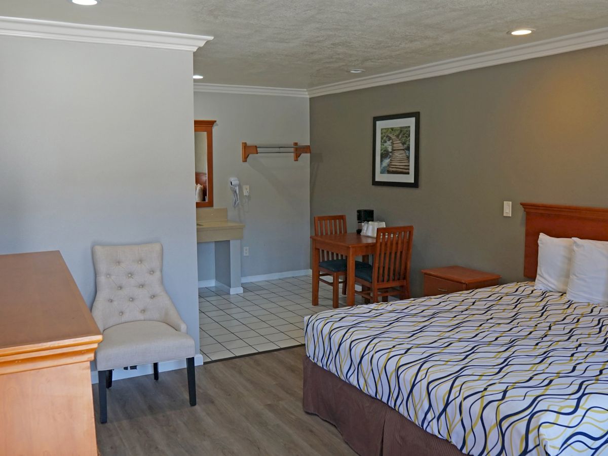 The image shows a hotel room with a bed, desk, chair, small dining set, and bathroom sink area. The room is decorated in neutral tones.