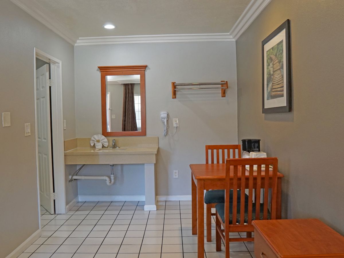 A room with a small dining table, two chairs, a sink area with a mirror, and a wall-mounted hairdryer. The walls are painted gray with framed art.