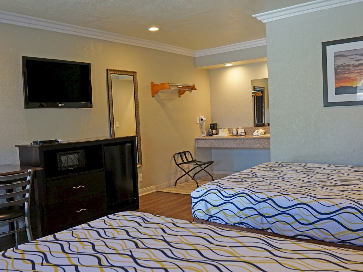 The image shows a hotel room with two beds, a TV, a table and chairs, a dresser, a mirror, and a painting on the wall. There is also a vanity area.