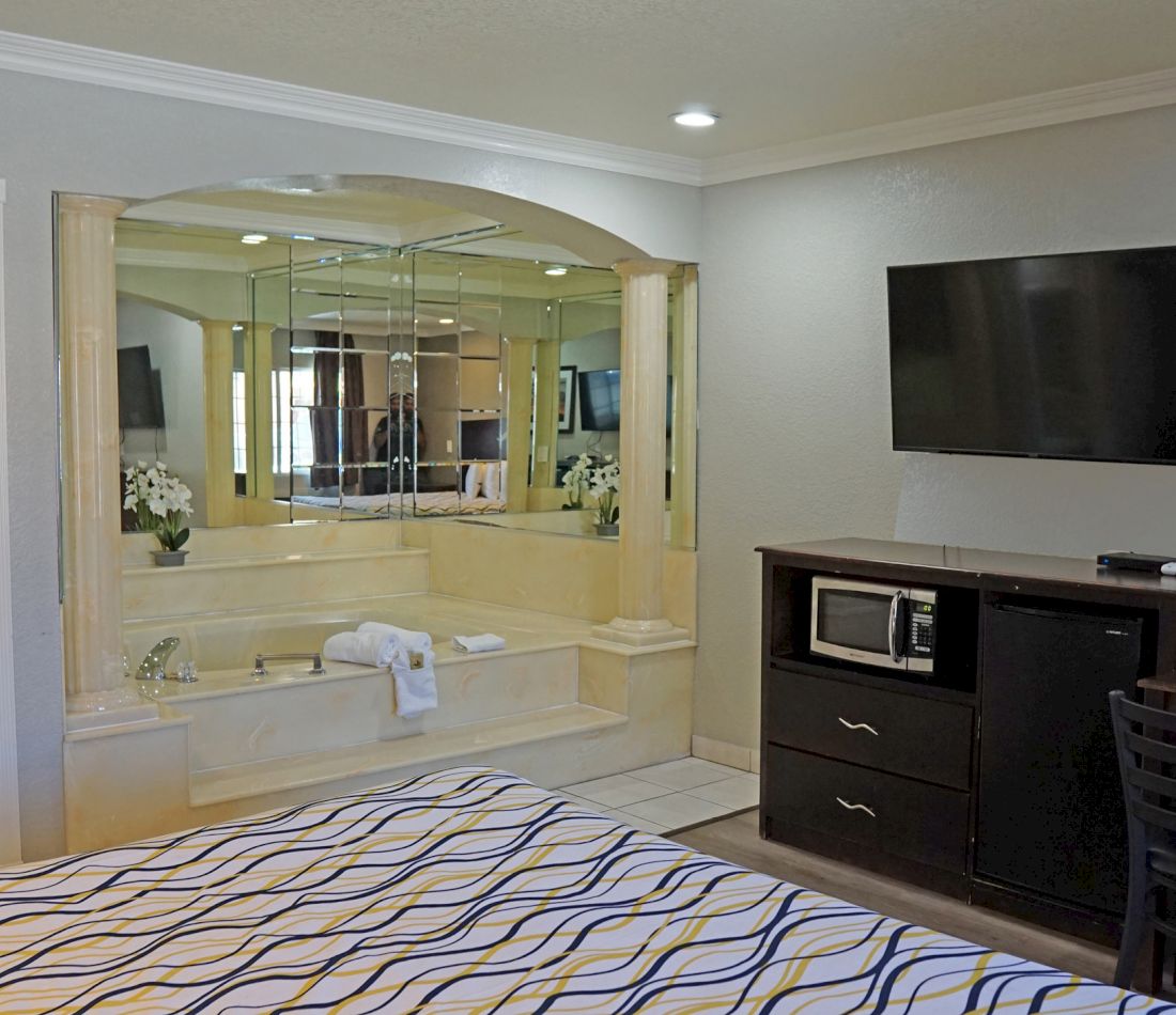 A hotel room with a large bed, a jacuzzi tub with a mirrored wall, and a flat-screen TV on a wooden cabinet.