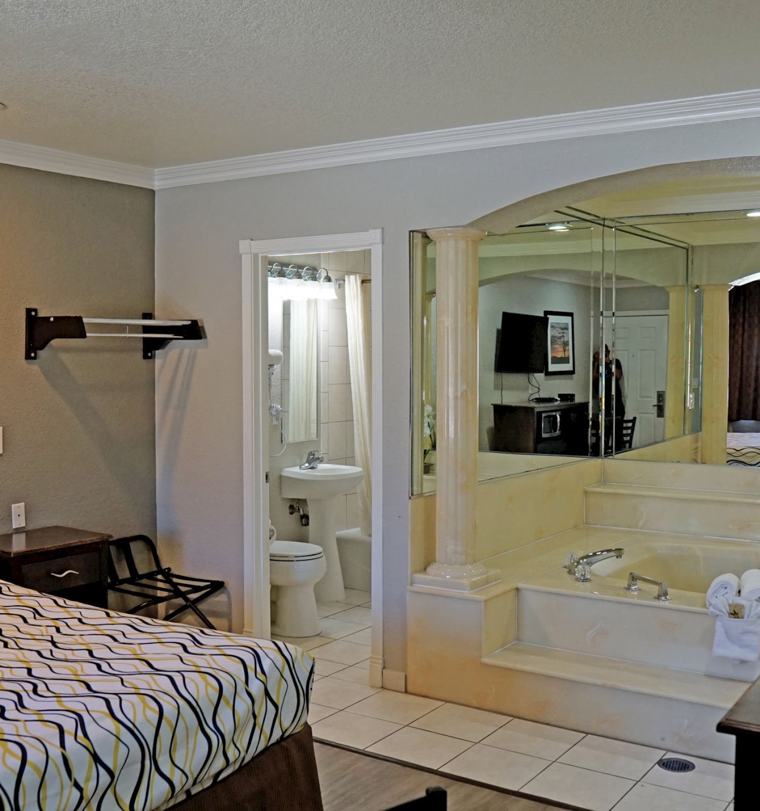 A hotel room with a bed, desk, TV, and an open bathroom featuring a spa tub next to the bed, along with a separate toilet area.