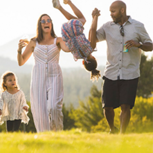 A family of four is enjoying a sunny day outdoors. The parents are playfully swinging one child by the arms while the other child walks alongside them.
