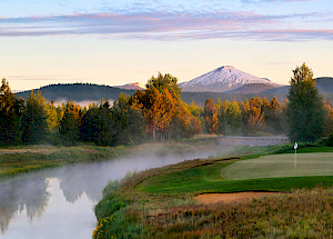 A beautiful golf course with a river and trees in the foreground, a snow-capped mountain in the distance, and a clear sky with light clouds ending the sentence.