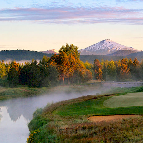 A beautiful golf course with a river and trees in the foreground, a snow-capped mountain in the distance, and a clear sky with light clouds ending the sentence.