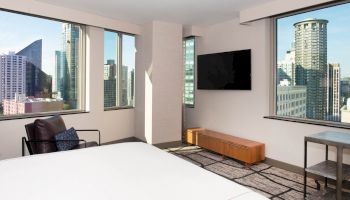 A modern hotel room with a bed, armchair, TV, and large windows showcasing a city skyline with various high-rise buildings.