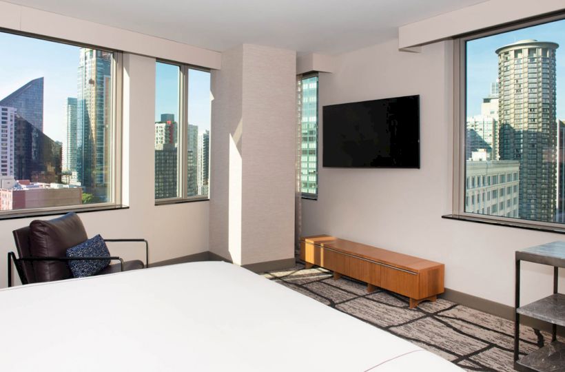 A modern hotel room with a bed, armchair, TV, and large windows showcasing a city skyline with various high-rise buildings.