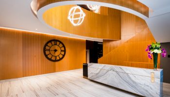 The image shows a modern reception area with a marble desk, wooden wall paneling, a large clock, and contemporary lighting fixtures.
