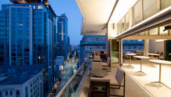 A modern rooftop terrace with outdoor seating overlooks a cityscape of tall buildings and a bustling street below, captured at dusk.