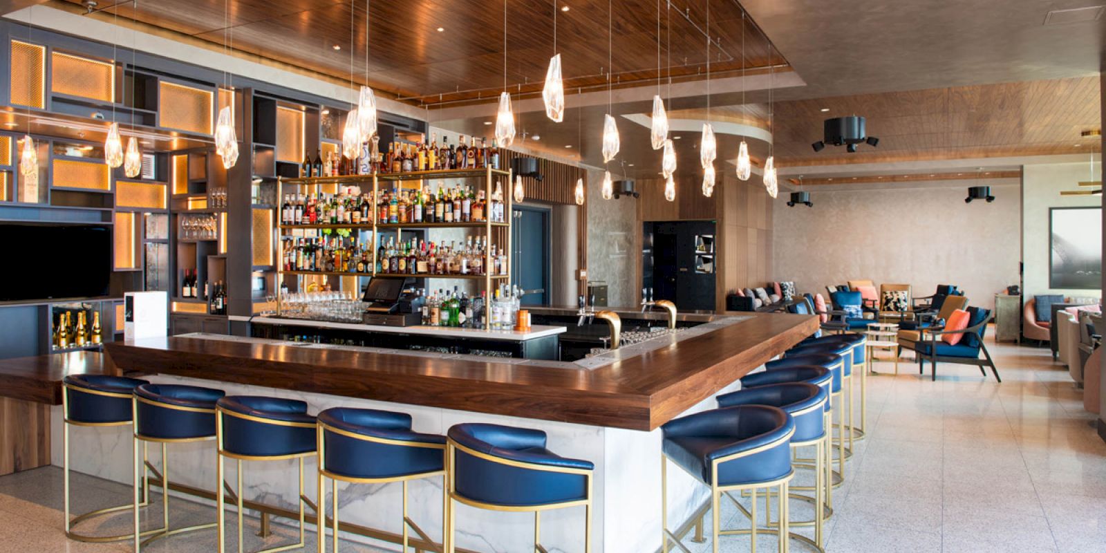 A modern bar with a wooden counter, blue upholstered bar stools, hanging lights, and a well-stocked liquor shelf in a stylish, open lounge area.