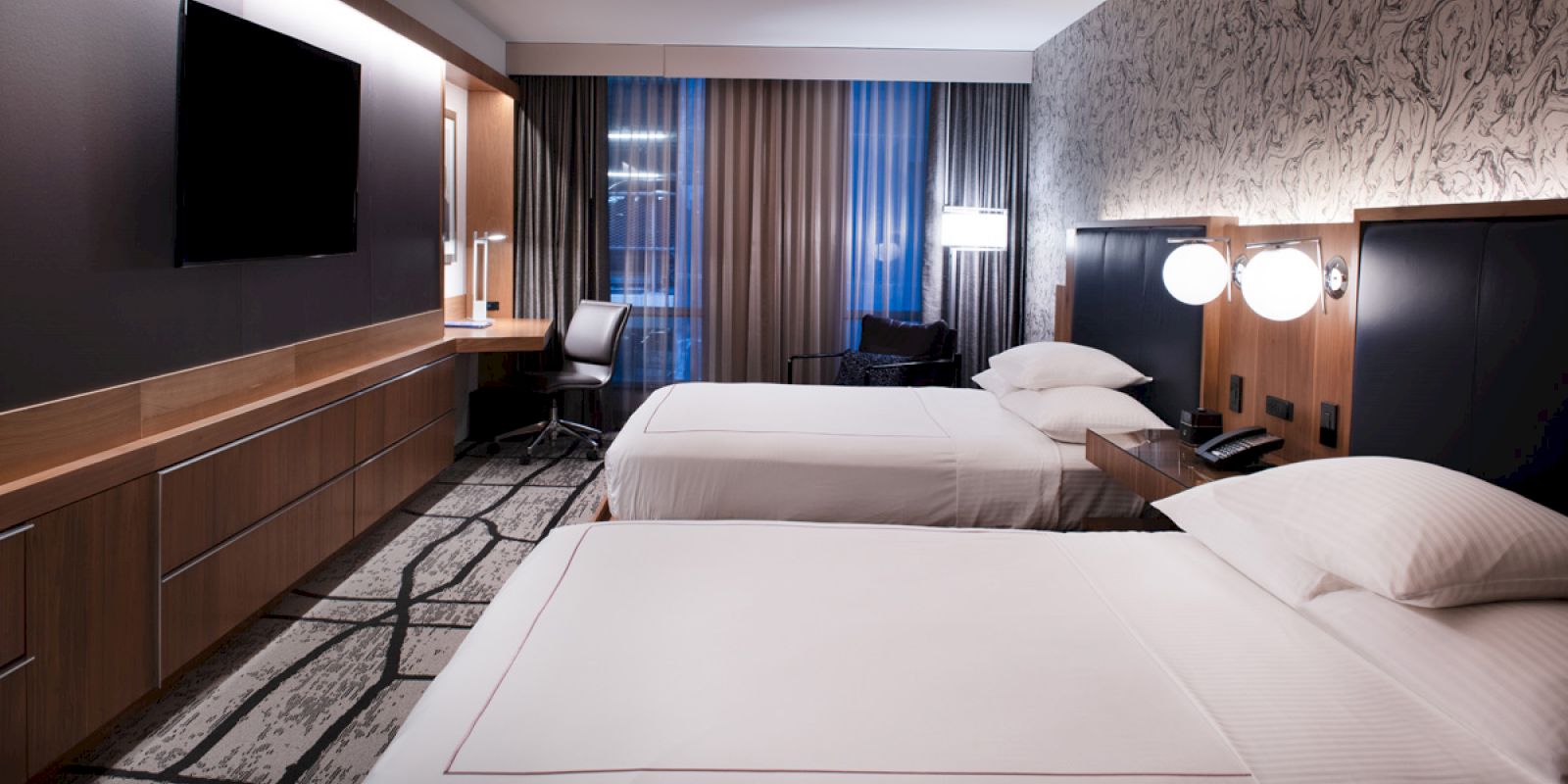 A modern hotel room with two neatly made beds, a flat-screen TV, a work desk, and stylish decor, featuring a patterned accent wall and large windows.