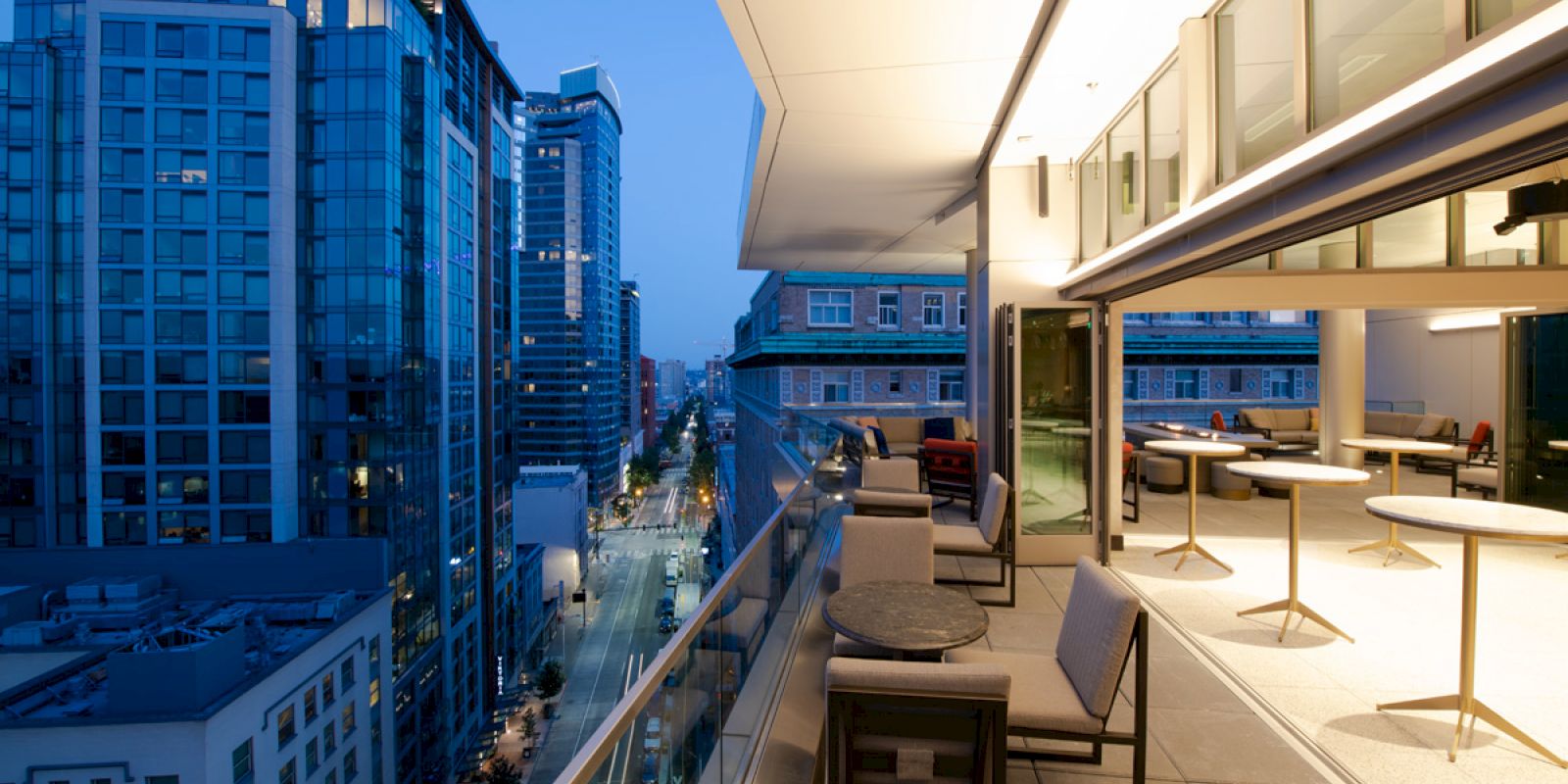 A modern cityscape with tall buildings at dusk. A spacious balcony with seating and tables overlooks the street below, providing a relaxing view.