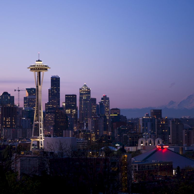 The image showcases the Seattle skyline at dusk, highlighted by the iconic Space Needle, with mountains and a mix of buildings in the background.