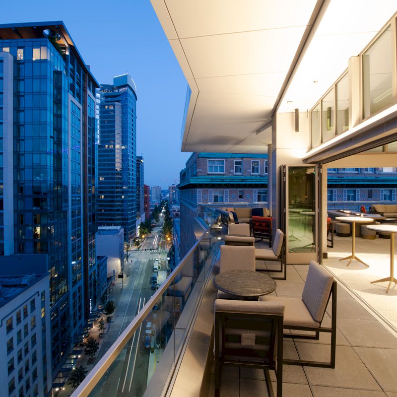 A modern cityscape at twilight viewed from a sleek outdoor terrace of a high-rise building with tables and chairs.