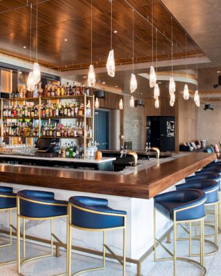 The image shows a modern bar with a variety of bottles on display, surrounded by blue barstools and featuring a sleek, contemporary design.