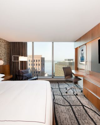 A modern hotel room features a bed, a desk with a chair, a flat-screen TV, and large windows offering a city view.