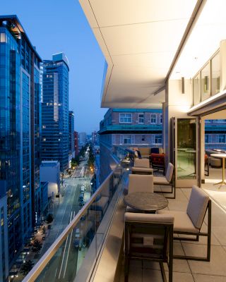 A modern balcony with chairs and tables overlooks a cityscape with tall buildings and lit streets at dusk, creating an inviting, urban atmosphere.