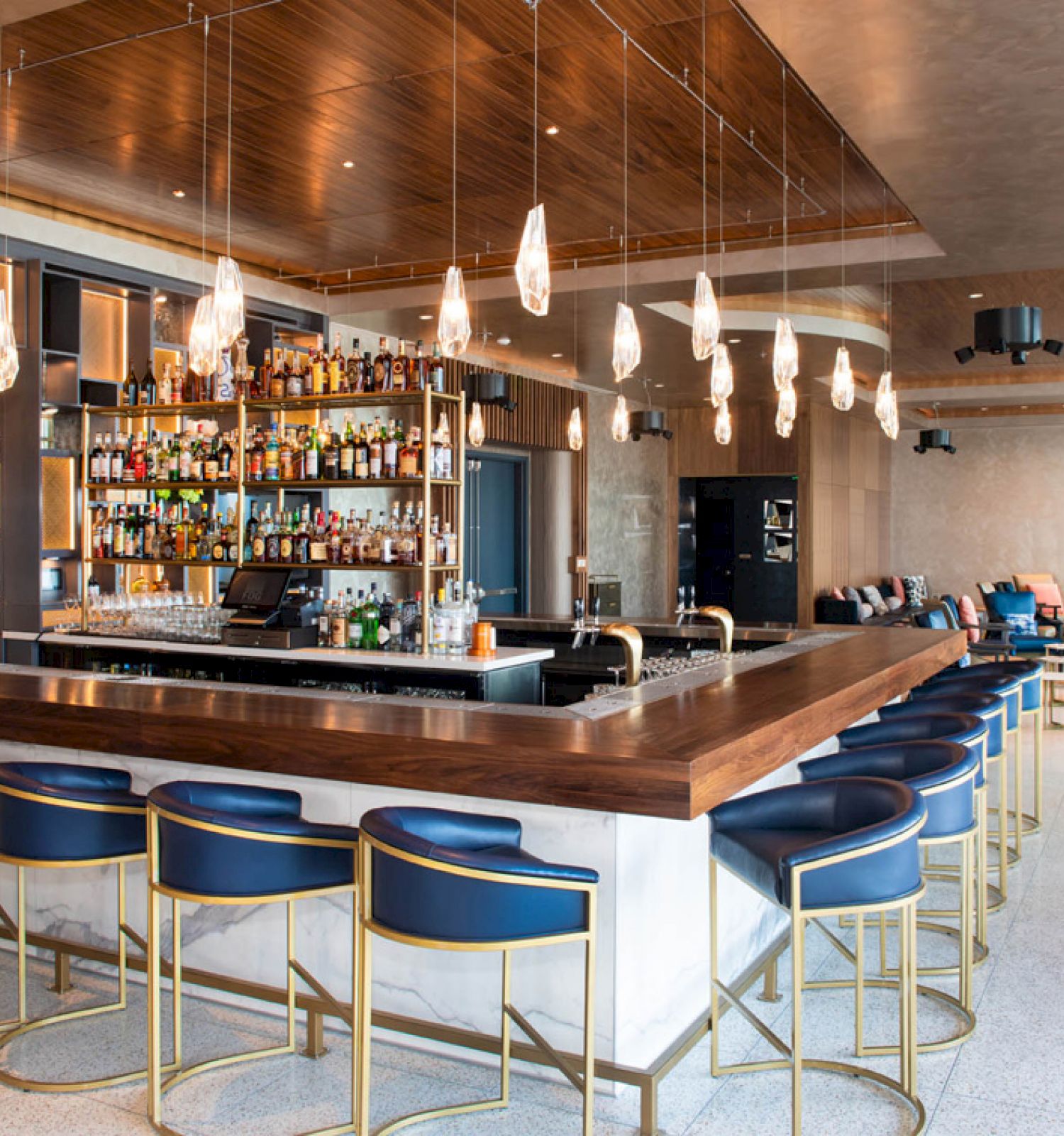 This image shows a modern bar area with a central counter, blue bar stools, hanging pendant lights, and a well-stocked liquor shelf.