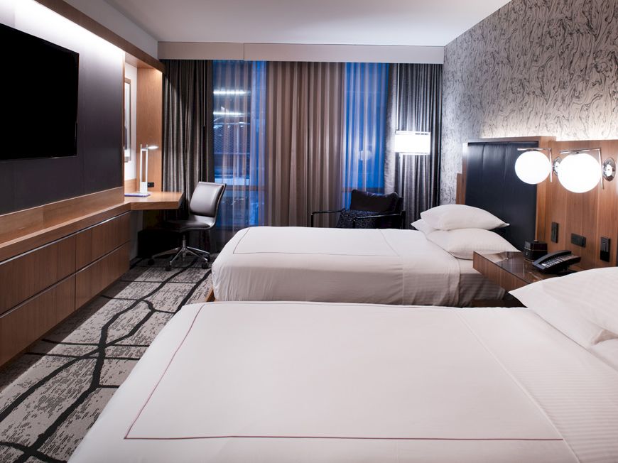 A modern hotel room with two neatly made beds, a wall-mounted TV, a desk with a chair, and warm lighting.