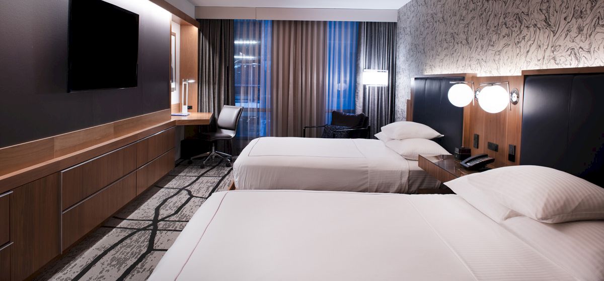 A modern hotel room with two neatly made beds, a wall-mounted TV, a desk with a chair, and warm lighting.