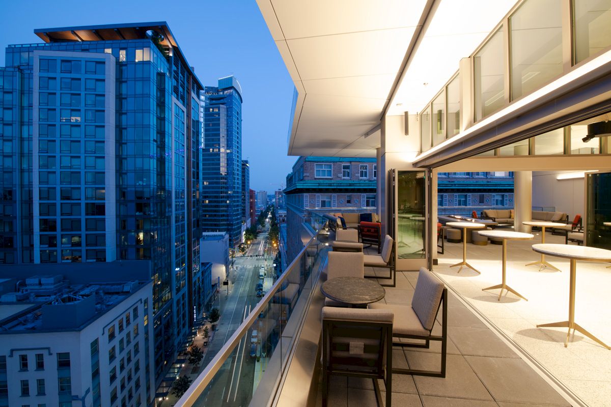 A modern balcony with tables and chairs overlooks a city street with tall buildings, captured during the evening.