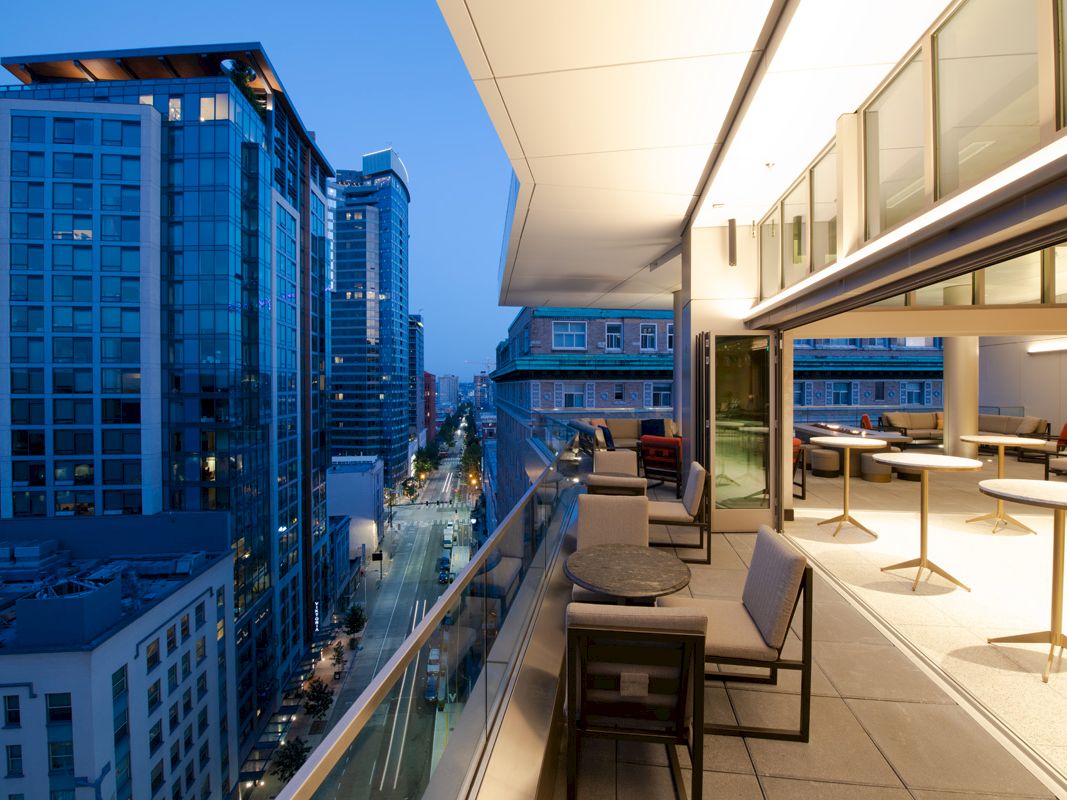 A modern balcony with tables and chairs overlooks a city street with tall buildings, captured during the evening.