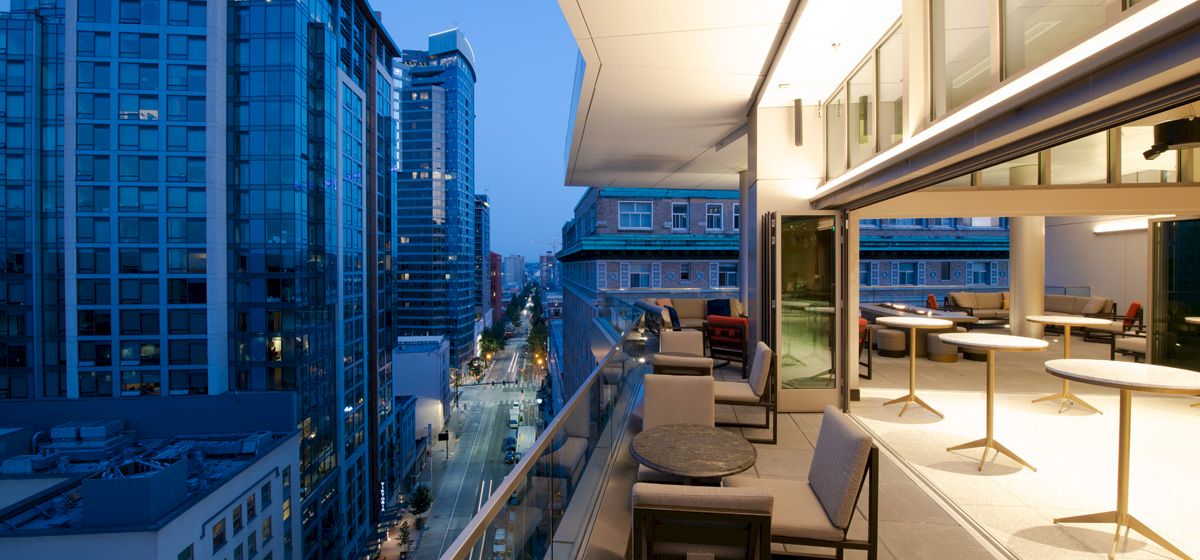 A modern balcony with tables and chairs overlooks a city street with tall buildings, captured during the evening.