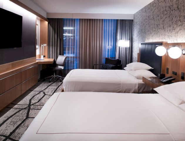 The image shows a modern hotel room with two beds, a TV, a work desk, and stylish decor with large windows and curtains.