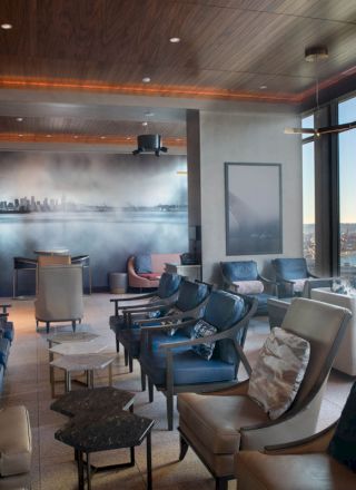 A stylish lounge with modern seating, large windows offering a scenic view, and artwork depicting a cityscape on the back wall, lit warmly.