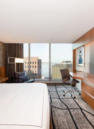 A modern hotel room features a large bed, desk, chair, wall-mounted TV, and a view of buildings outside the window.