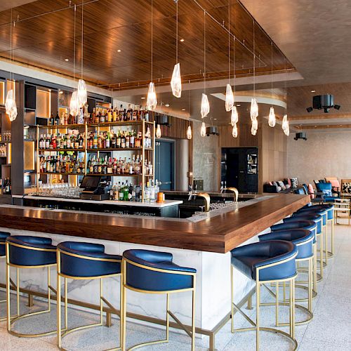 This image shows a modern bar with a row of blue bar stools, a well-stocked liquor shelf, stylish lighting, and a cozy seating area in the background.