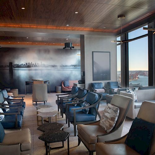 The image shows a modern lounge with leather chairs, a view of the ocean through large windows, and artwork on the back wall.