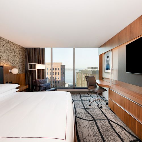 A modern hotel room with a large bed, wall-mounted TV, desk, office chair, and floor-to-ceiling window offering a city view and some buildings outside.