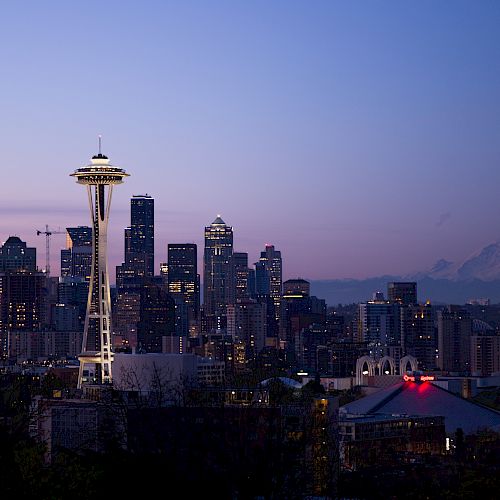 A cityscape view featuring the iconic Space Needle in Seattle at twilight, with buildings and distant mountains visible in the background.
