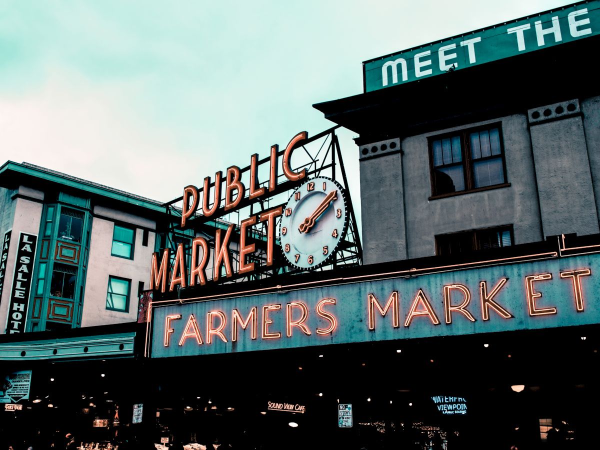 A well-known public market with a neon "FARMERS MARKET" sign and a clock above. Buildings around show signage, including a hotel and "MEET THE" phrase.