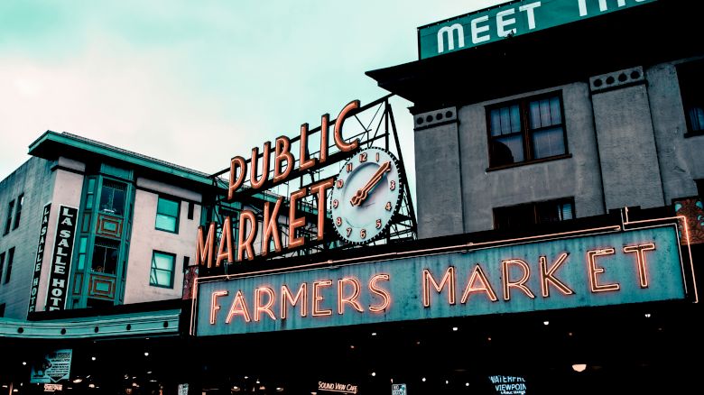 A well-known public market with a neon "FARMERS MARKET" sign and a clock above. Buildings around show signage, including a hotel and "MEET THE" phrase.
