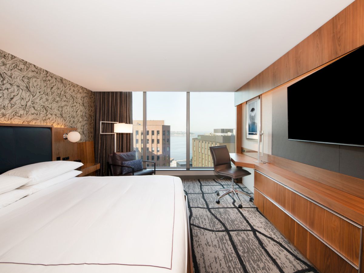 A modern hotel room with a large bed, wall-mounted TV, desk, chair, and a view of city buildings through a large window, ends the sentence.