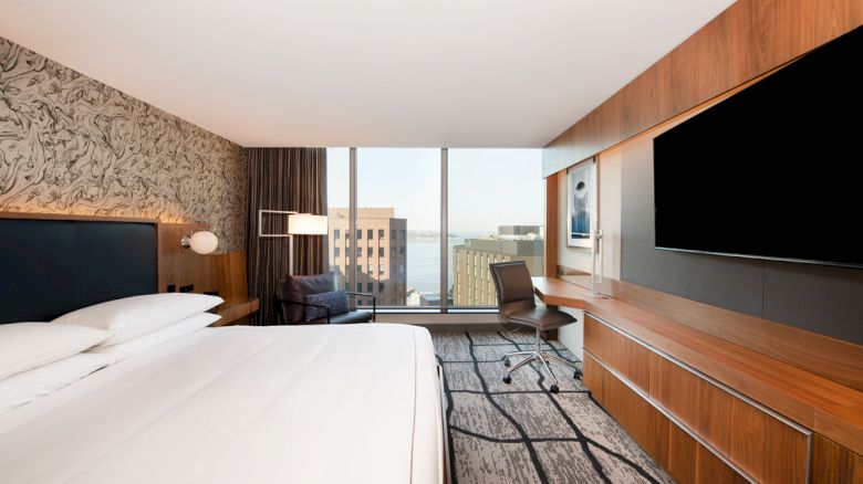 A modern hotel room with a large bed, wall-mounted TV, desk, chair, and a view of city buildings through a large window, ends the sentence.