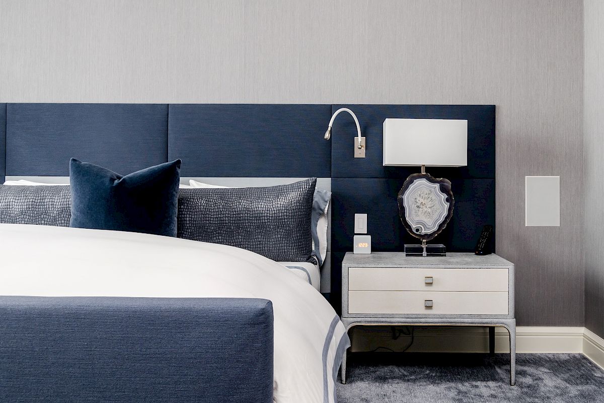 A modern bedroom with a blue and white color scheme, featuring a blue headboard, a white nightstand, and a lamp on top, beside the bed ending the sentence.