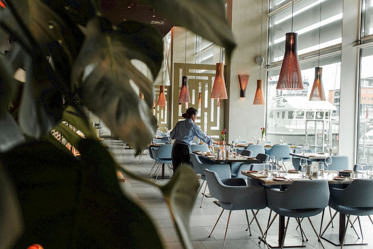 A person is setting up or arranging tables in a modern, stylishly decorated restaurant with large windows and hanging lights.