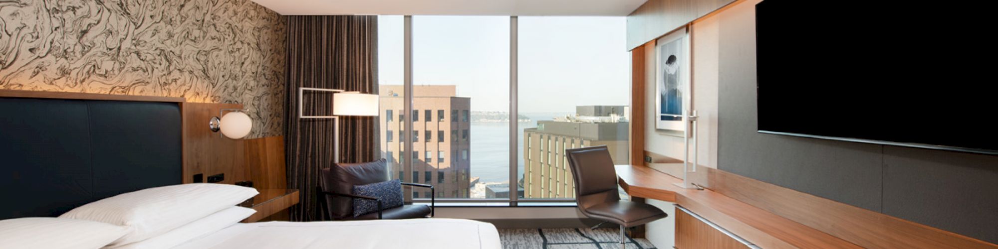 A modern hotel room features a large bed, wall-mounted TV, desk, chair, and a window with a cityscape view, along with stylish décor on the walls.
