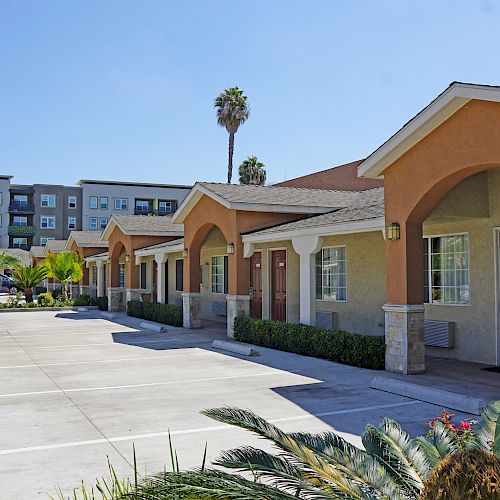 The image shows a row of single-story buildings, possibly a motel or apartment complex, with an empty parking lot and a multi-story building in the background.