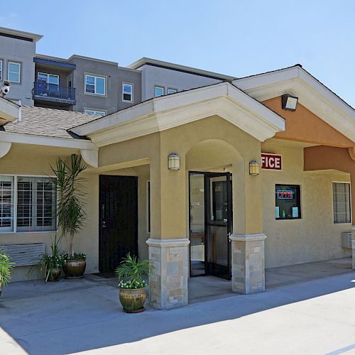 This image shows the exterior of a building with a sign indicating it is an 'office,' featuring plants near the entrance and palm trees in the background.