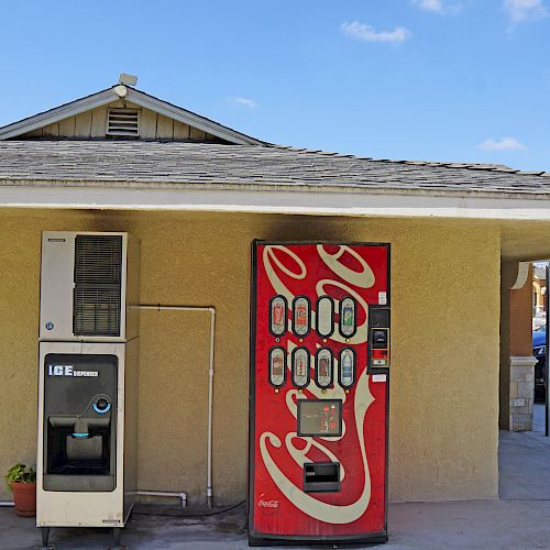 The image shows a vending machine area with a soda machine and a water dispenser under a small shelter next to a parking lot.