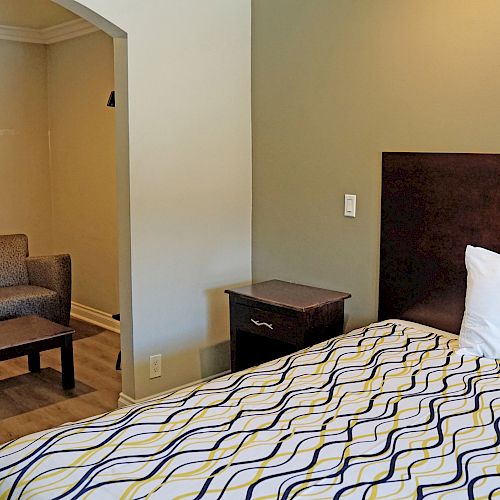 This image shows a well-lit hotel room with a bed, side table, framed art, and sitting area with two chairs and a coffee table.