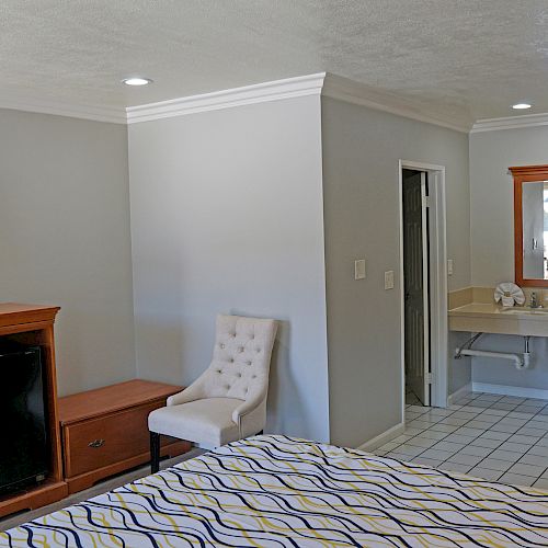 The image shows a tidy hotel room with a bed, TV, dresser, armchair, table, mirror, sink area, and a wall-mounted hairdryer.