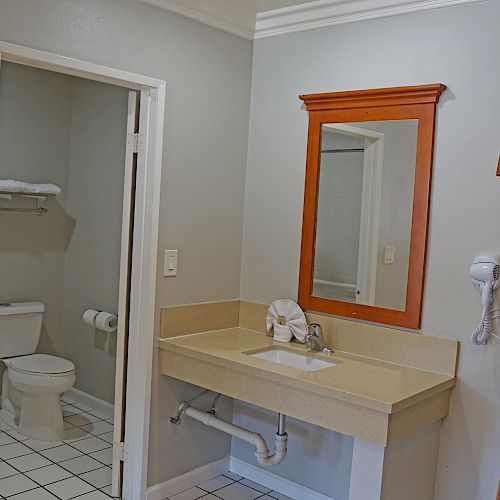 The image shows a bathroom with a sink, mirror, wall-mounted hairdryer, towel rack, and toilet with neatly rolled towels on a shelf.