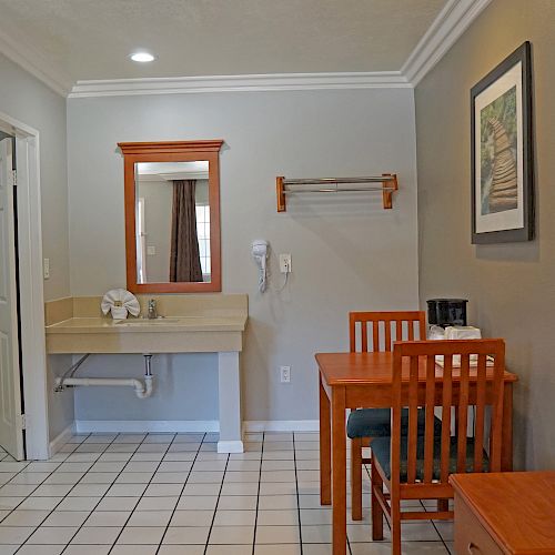 A room with a sink, mirror, towel rack, table with chairs, a picture on the wall, and a small wooden cabinet on a tiled floor.